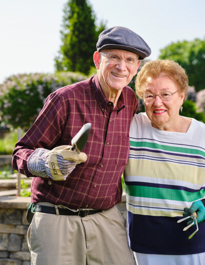 Happy older couple wears gloves while holding small tools, and smiles at the camera while gardening.