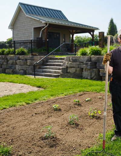 Retiree holds a metal tined rake, wearing gloves and a baseball cap while gardening at the Quarryville Life Plan community.