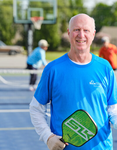 Retiree in a blue Quarryville Retirement Community t-shirt holds a ball and raquet on the tennis court.