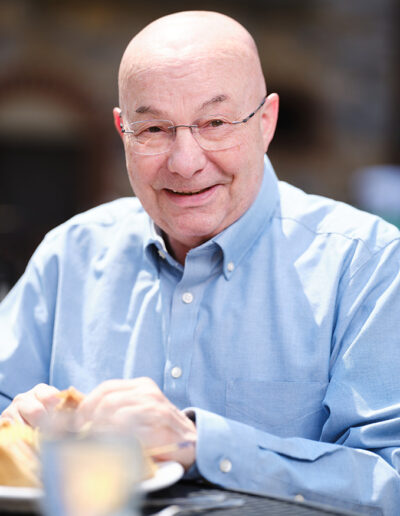 older man smiling in a blue shirt with a meal, at his life plan community, enjoying retirement.