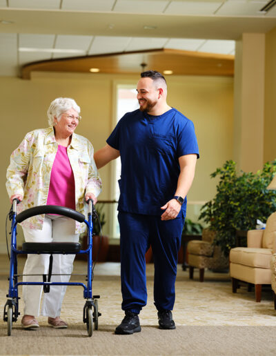 Skilled staff in nursing blues walks with a woman using a mobility assistant device.