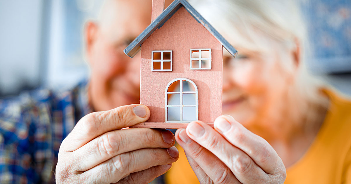 Retired couple blurred in the background of a small home they are holding in front of themselves, a visual of looking into daily life at a life plan community.