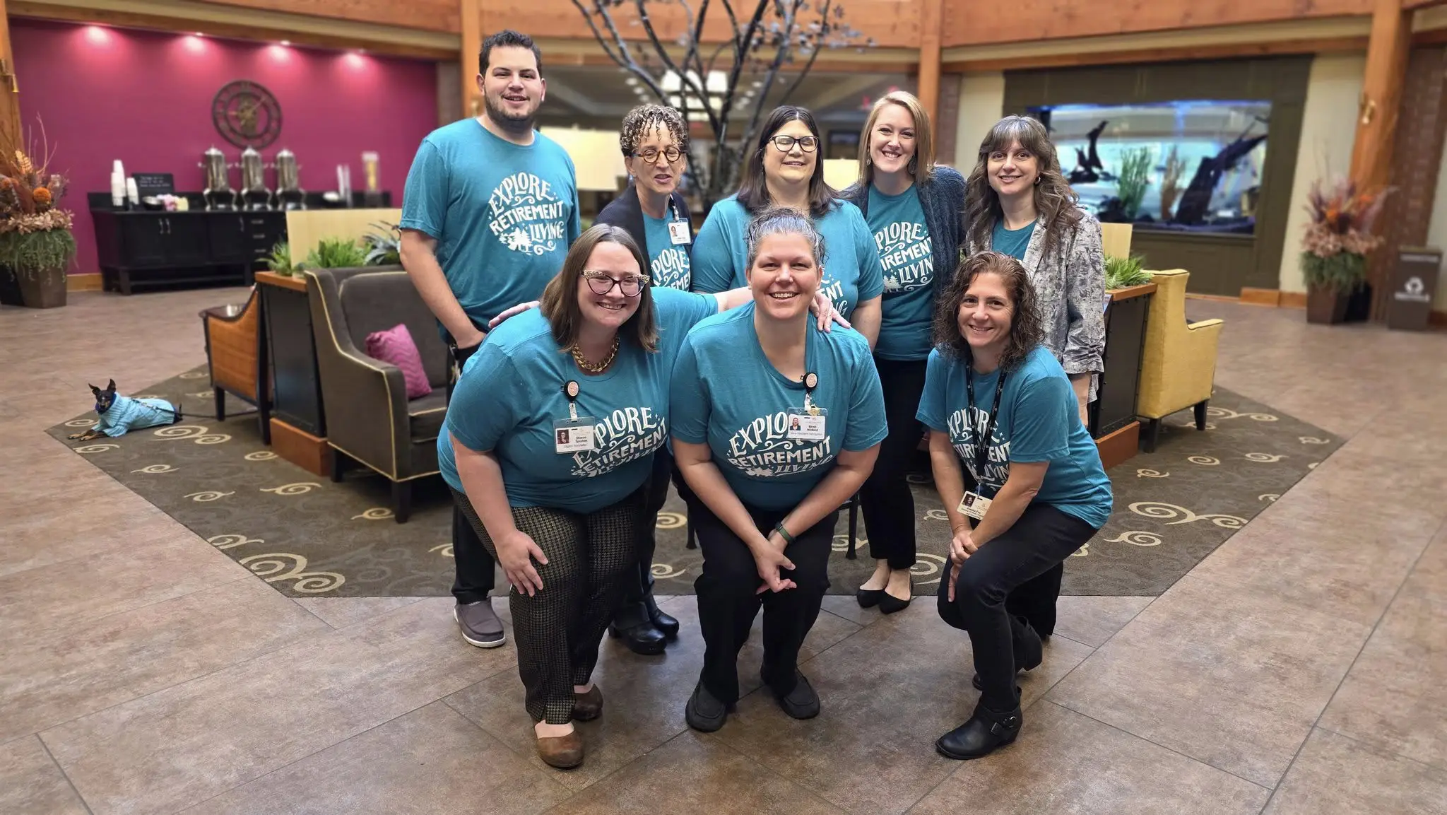 Eight people in teal Explore the excitement t-shirts pose and smile inside a spacious, well-lit lobby with colorful couches, plants, and a modern decor.