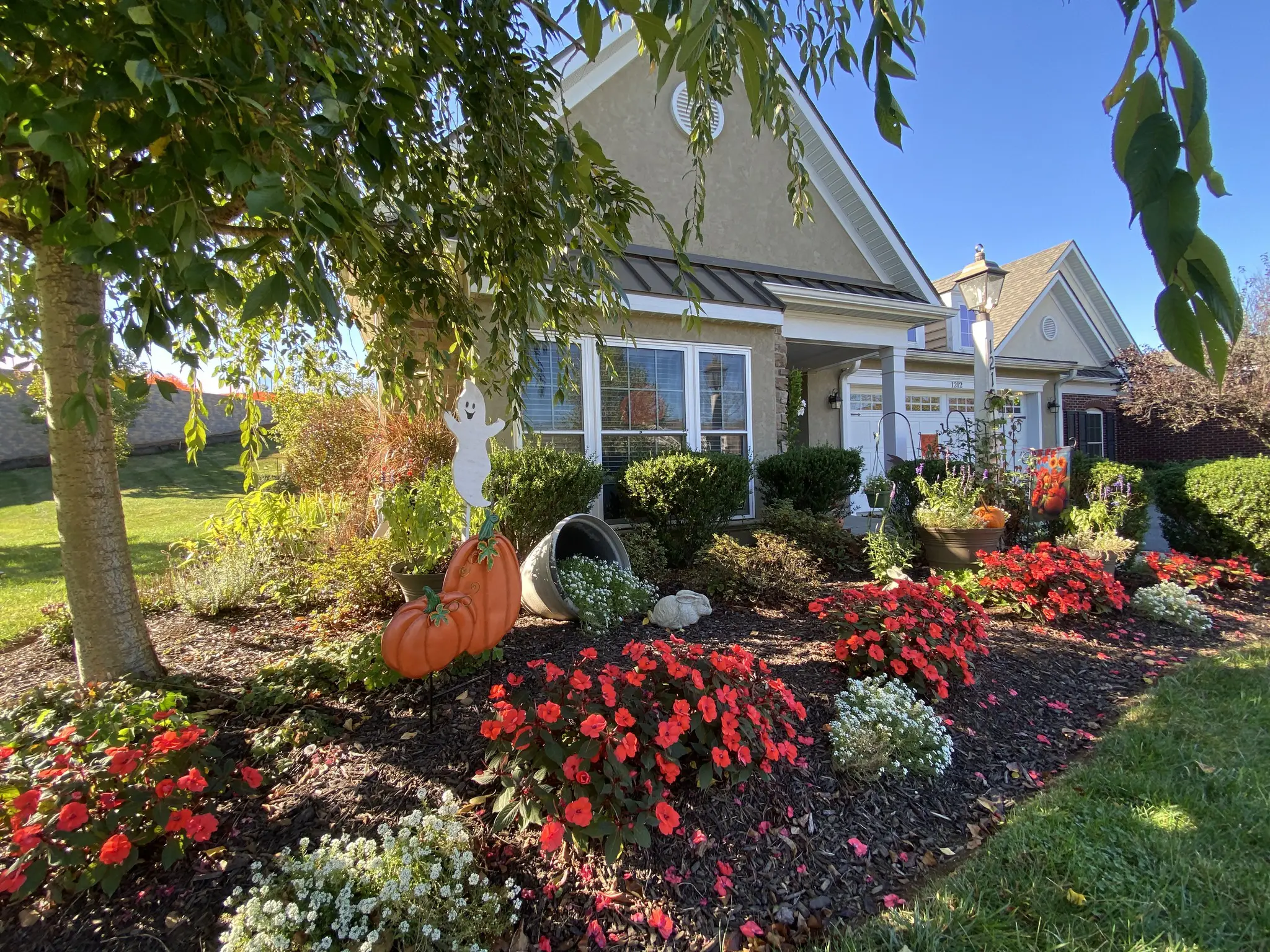 A house with a well-kept garden, red flowers, a pumpkin and ghost Halloween decoration, and a cornucopia. The sky is clear and blue.